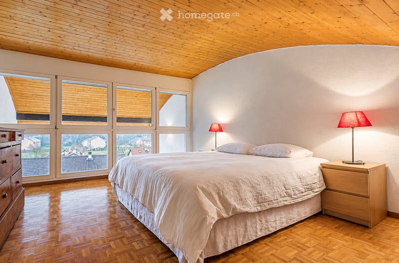 Bright bedroom with wooden flooring, large windows, a neatly made bed, nightstands with lamps, and a wooden dresser.