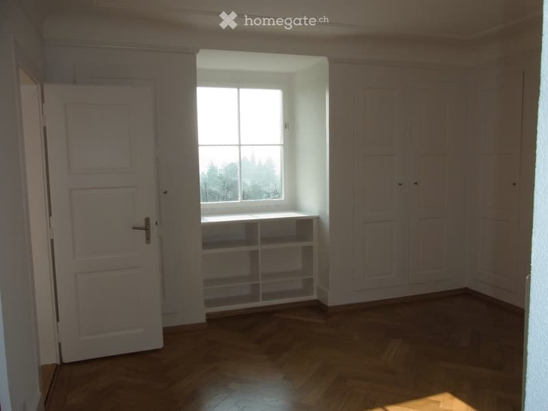 Empty room with wooden flooring, white walls, built-in shelving unit, and a window with a view of trees and a church spire.