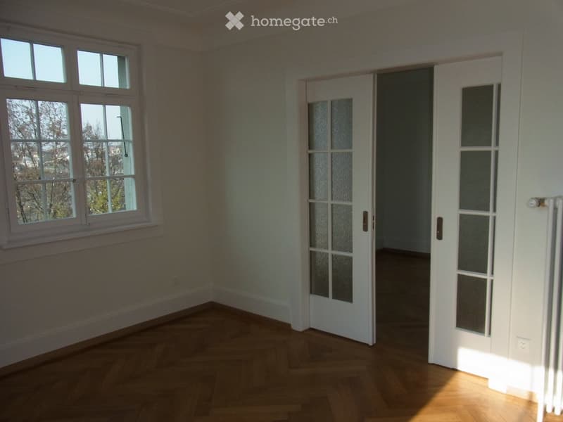 Empty room with wooden flooring, white walls, large windows, and a white door leading to another space.