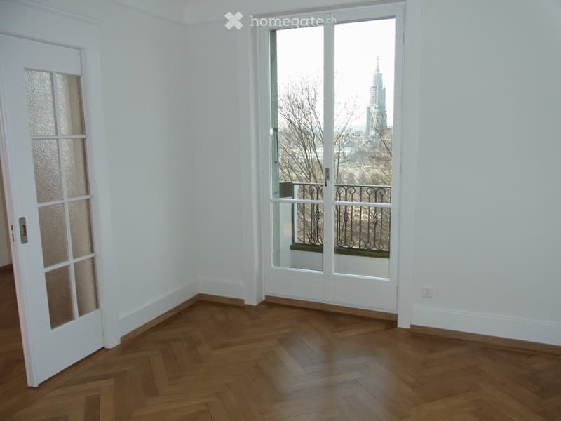 Empty room with wooden herringbone flooring, white walls, and a large window offering a cityscape view.