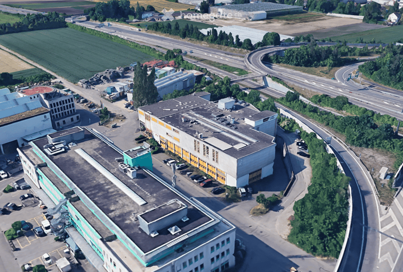 Industrial buildings in a suburban area, surrounded by trees and vegetation, featuring multiple parking areas and access roads.