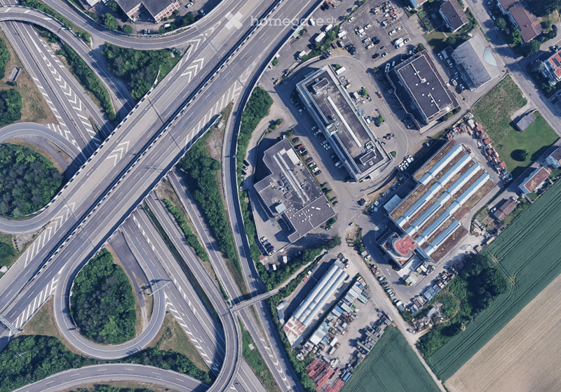 Highway with multiple lanes, buildings on the side, parking lot with several cars, trees, and plants, fields
