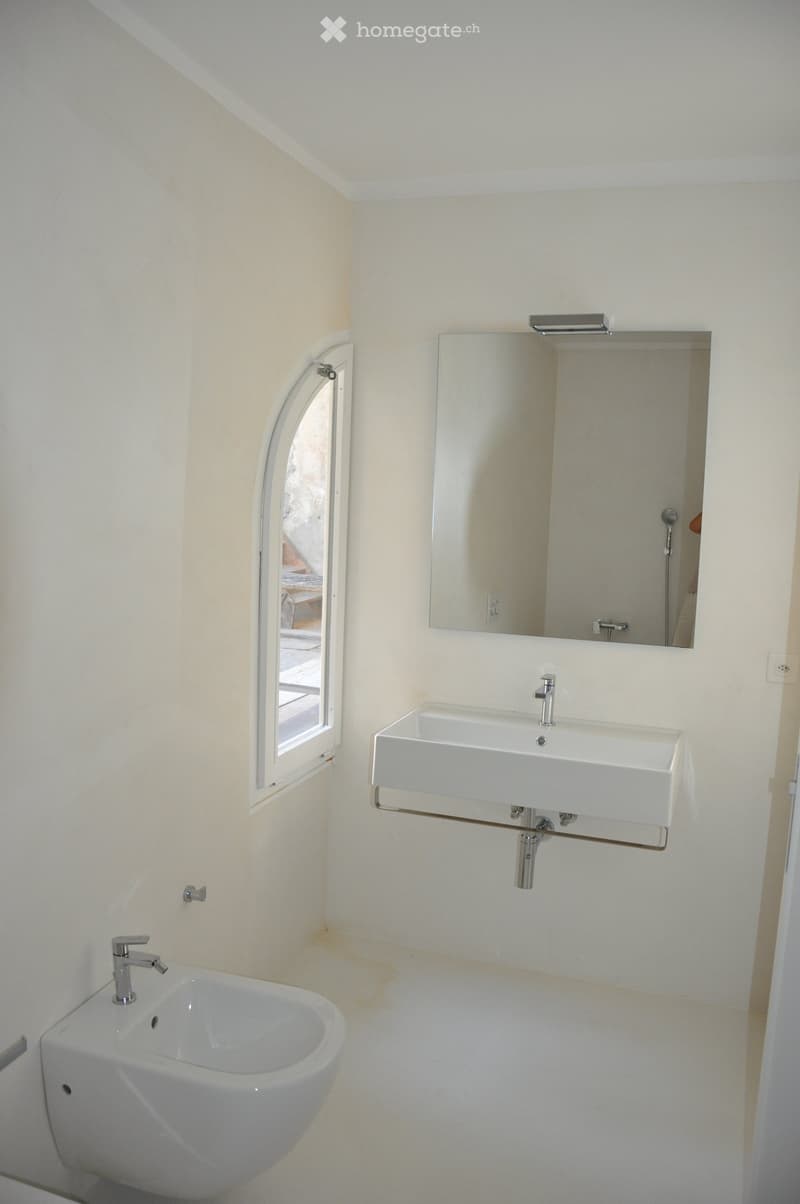 Modern bathroom with white walls, a large mirror, a sink with a modern faucet, and a bathtub. A small window with a curved glass panel is visible on the left side.
