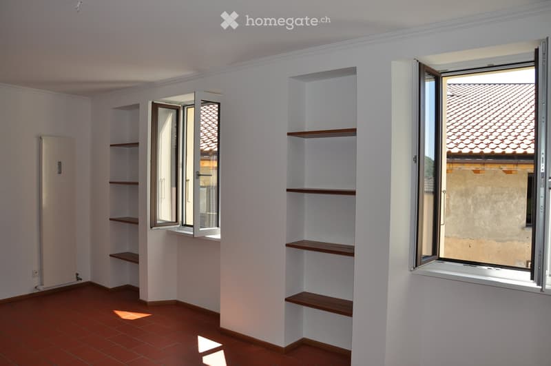 Empty room with white walls, wooden shelves, and large windows offering views of a tiled roof. The floor is made of terracotta tiles.