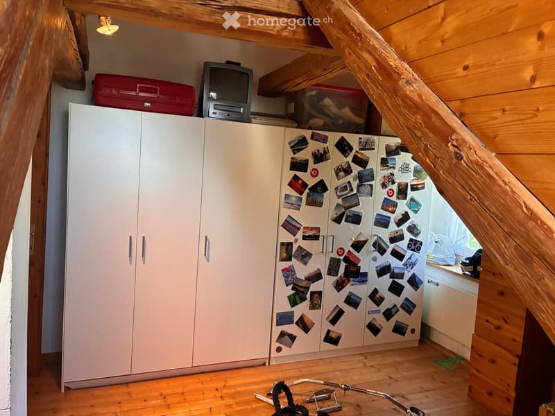 Attic room with wooden beams, white cabinet, refrigerator covered in photos, television, and a suitcase on top.