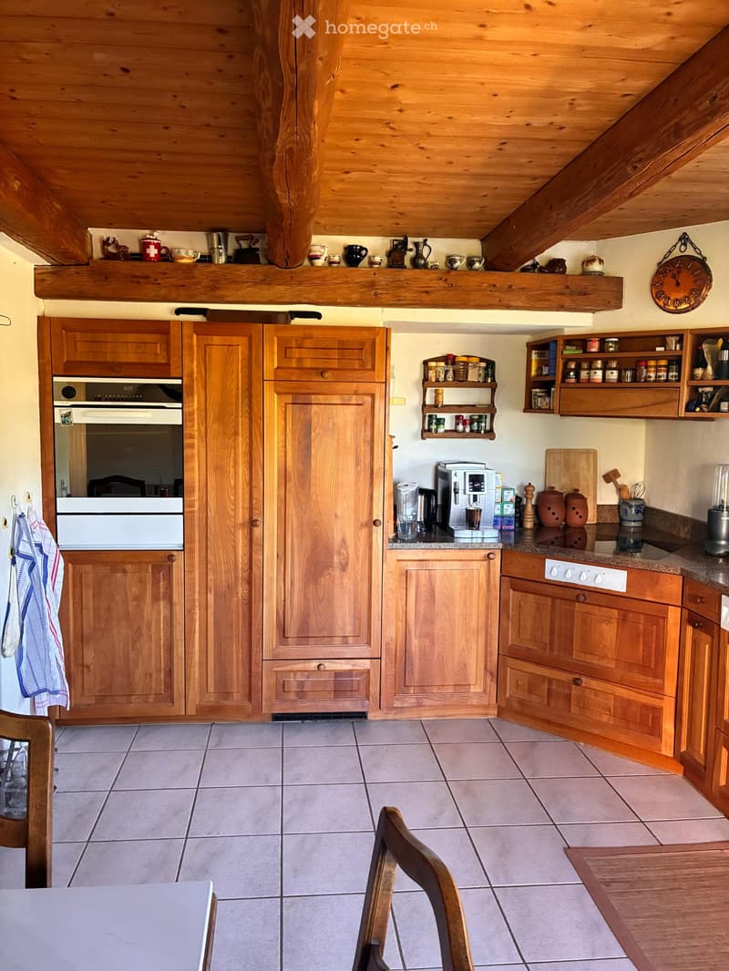 Cozy kitchen with wooden cabinets, tiled floor, and various appliances including an oven, coffee maker, and blender. Shelves hold spices and decorative items, and a dining area with a table and chairs is visible.