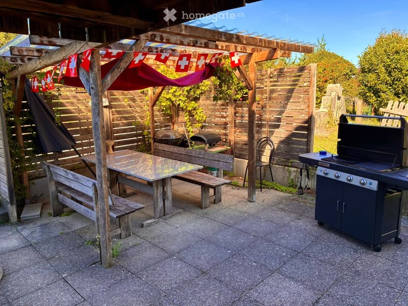 Wooden pergola with a table, benches, chairs, and a grill, surrounded by a wooden fence and greenery.