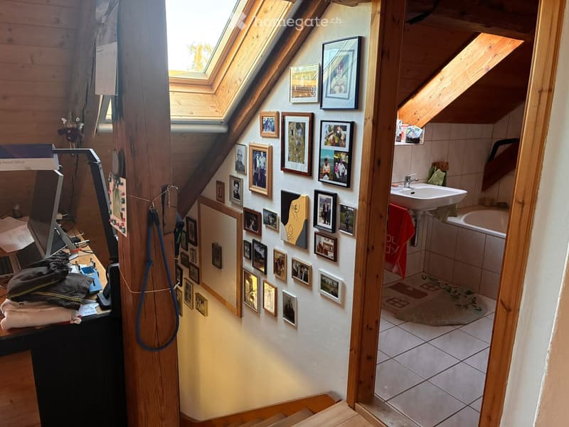 Loft-style room with wooden beams, skylight windows, and a small bathroom area visible through an open doorway. The room features a desk with a chair, framed pictures on the wall, and a towel hanging on the side.
