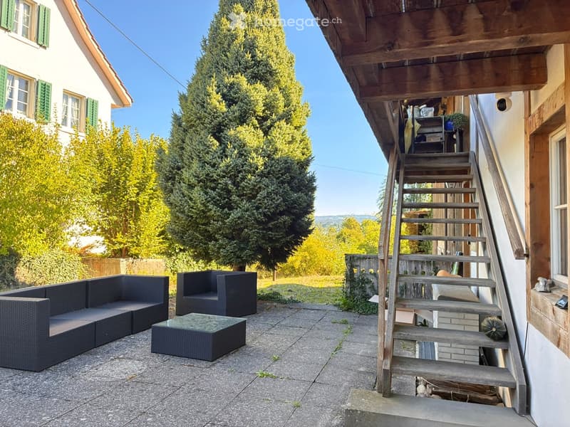 Outdoor patio with a black wicker sofa, matching armchair, and a glass-top coffee table. Wooden stairs lead up to a wooden deck area with additional seating and storage. The background features a house with green shutters and a lush, green landscape.