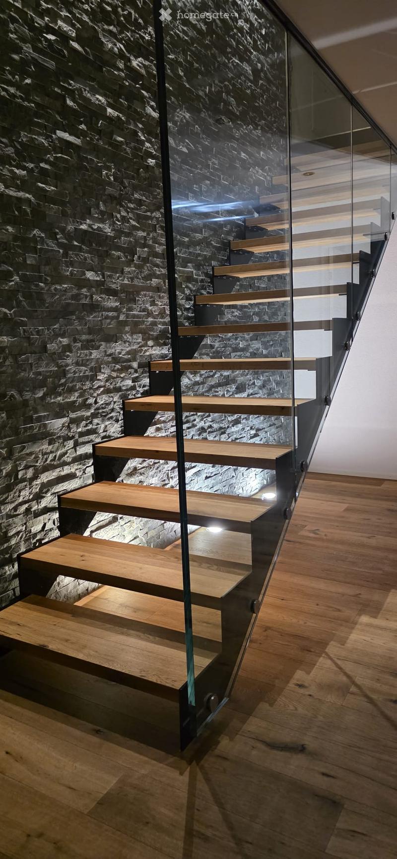 Modern staircase with wooden steps and glass railing, set against a textured stone wall, leading to an upper level with wooden flooring.