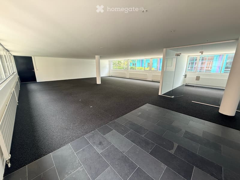 Empty room with dark carpet and tiled section, large windows, glass partition, and white walls.