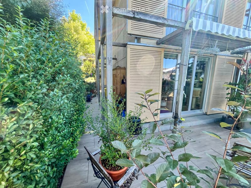 A narrow garden area with potted plants, a wooden bench, and a view of a modern house with large windows and a balcony.