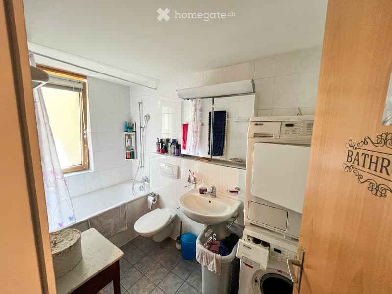 Bathroom with a bathtub, toilet, sink, mirror, washing machine, and various toiletries. The room features tiled walls and a window with a curtain.