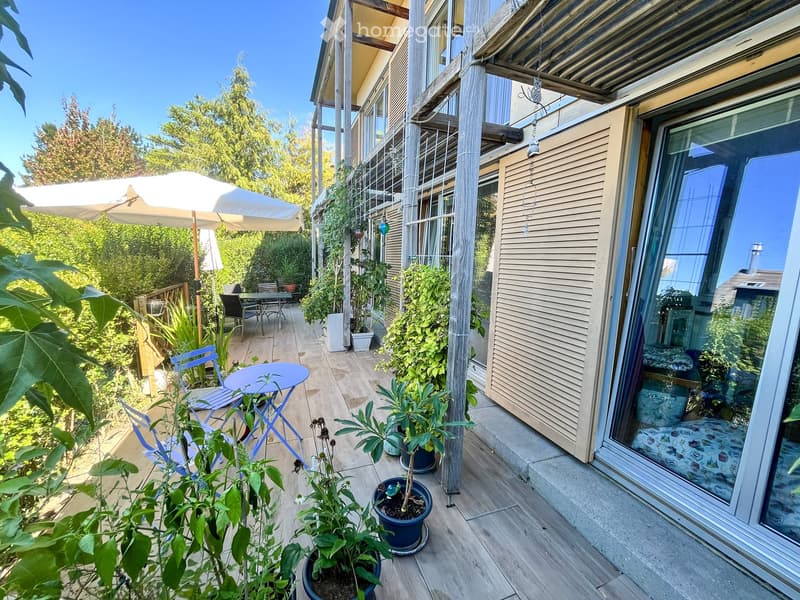 Balcony with wooden decking, potted plants, a small table, chairs, and an umbrella. Large sliding glass door leads to the interior.