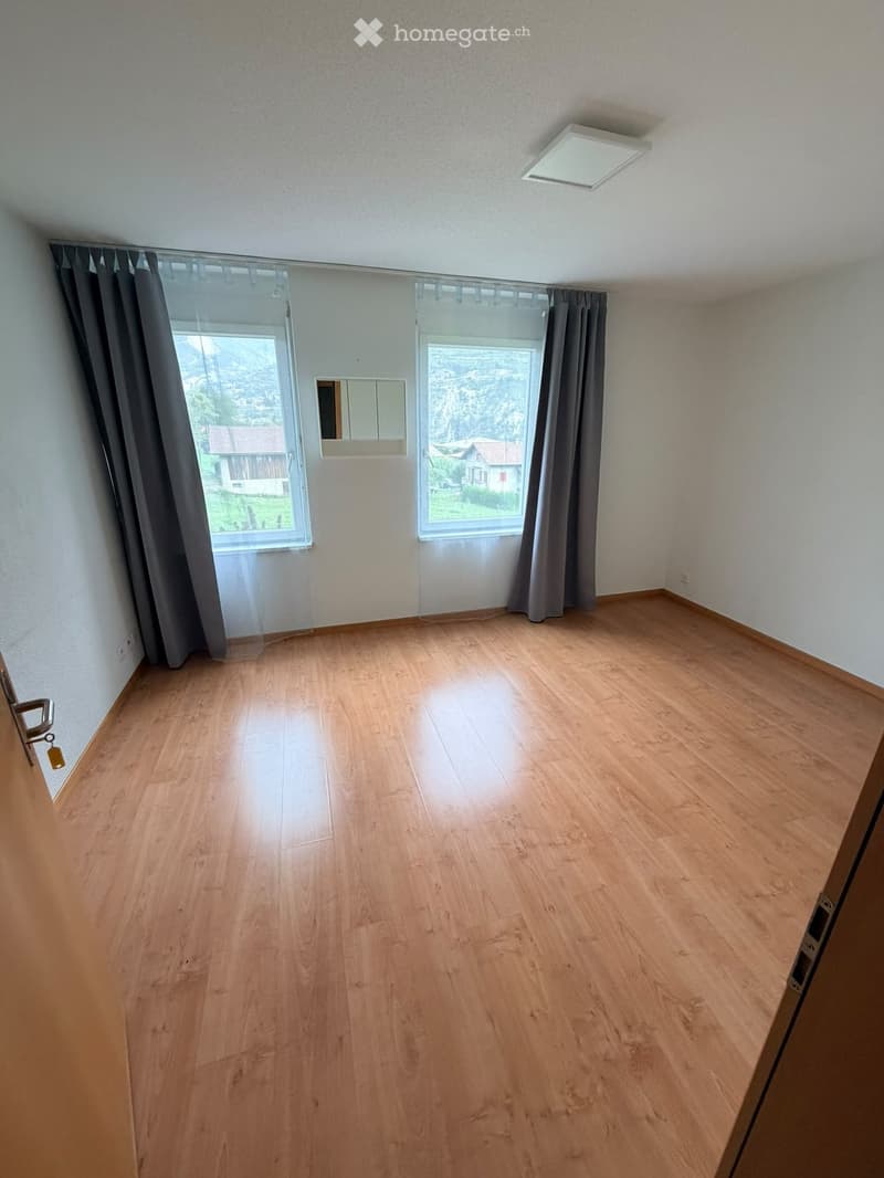 Empty room with wooden flooring, large windows with curtains, and a small mirror on the wall.