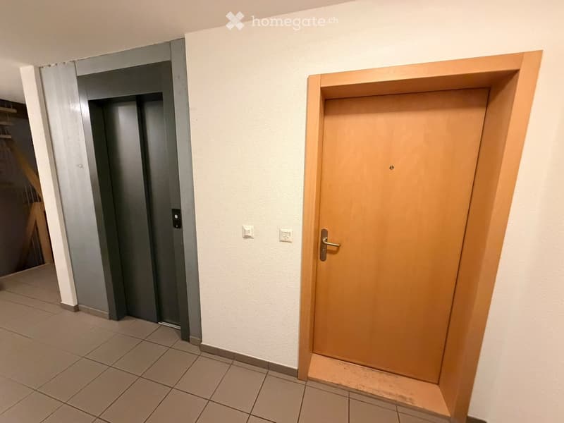 A hallway with tiled flooring, white walls, and two doors: a wooden door and a grey elevator door.
