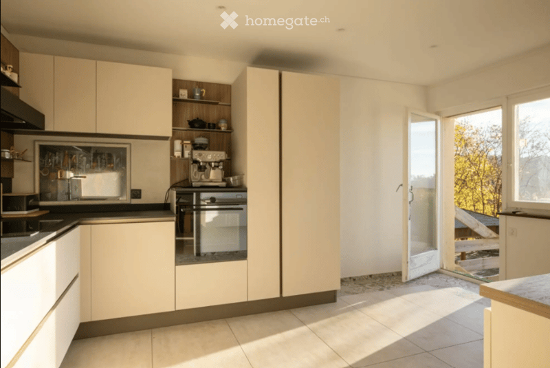 Modern kitchen with light-colored cabinets, stainless steel appliances, and a large window letting in natural light. The space includes a coffee maker, fridge, and sink.