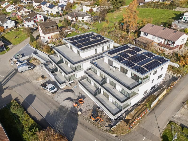 Multi-story apartment building with solar panels on the roofs, glass facades, surrounded by residential homes, green fields, and a road.