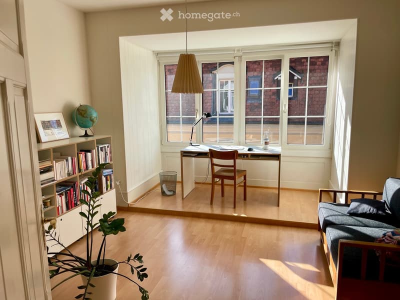 Bright, open living space with wooden flooring, large windows, a sofa, a desk with a chair, a bookshelf, a globe, and a potted plant.