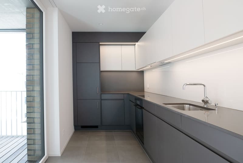 Modern kitchen with white and grey cabinets, a stainless steel sink, and a sleek faucet. The space features a large window with a view of a brick wall and balcony.