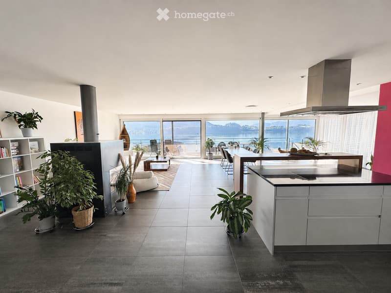 Open-plan living and kitchen area with large windows offering a scenic view, featuring a sofa, coffee table, kitchen island, bookshelf, and potted plants.