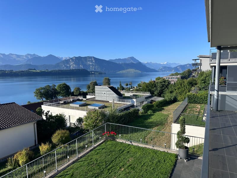 Garden area with a view of a lake and mountains, featuring a small lawn, potted plants, and a modern building with a balcony.