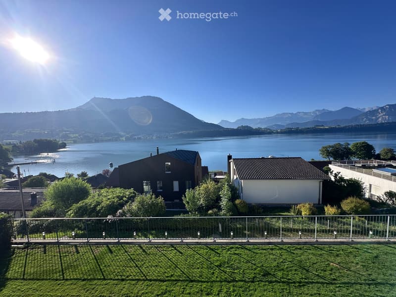 View of a serene lake and mountain range from a balcony or terrace with a metal railing and well-maintained lawn.