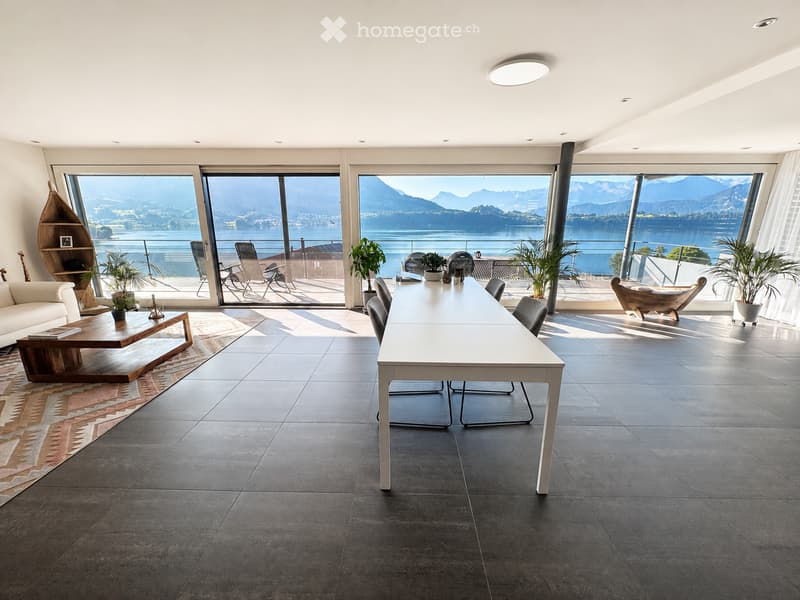 Modern open-plan living and dining area with large windows offering a scenic mountain view, featuring a white dining table, grey chairs, a white sofa, a wooden coffee table, and several potted plants.