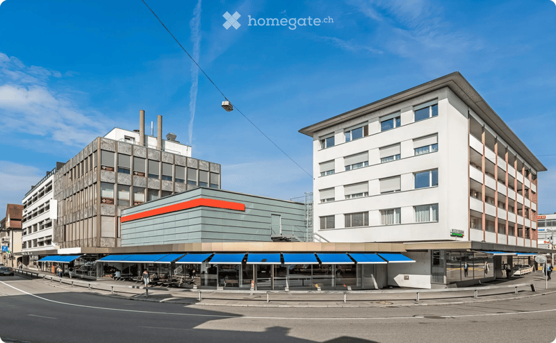 A multi-story building with a modern facade, featuring a mix of glass and concrete elements, with a restaurant area and seating under blue awnings.