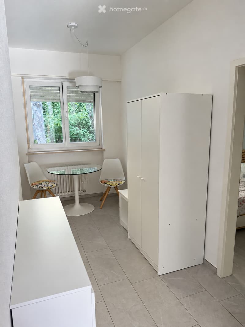 A small, modern room with a white tiled floor, a large window with blinds, a white pendant light, a round glass table with two colorful chairs, and a white cabinet.