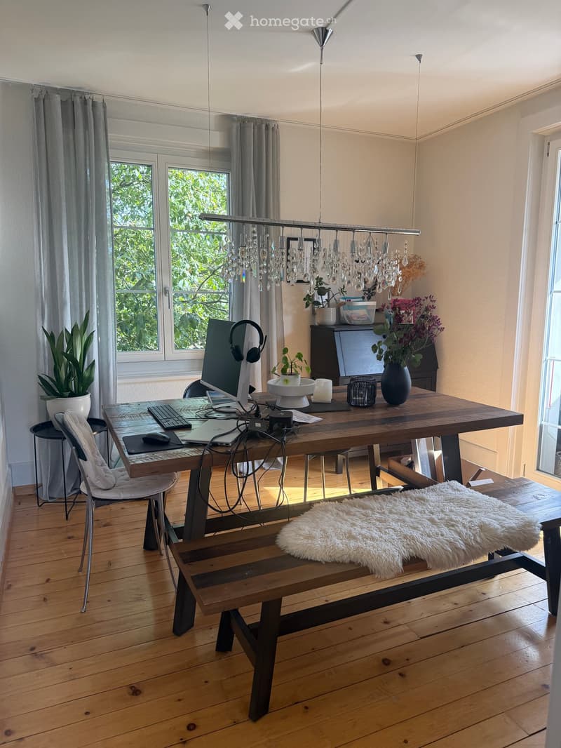 A well-lit room with wooden flooring, a large wooden table with a laptop, keyboard, mouse, and headphones, a bench with a fur throw, and a chandelier hanging from the ceiling. There are plants and a vase on the table, and large windows with curtains.