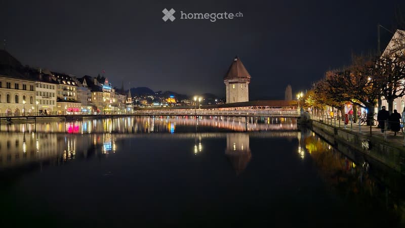 City by the river with streetlights and bridges, historic tower, buildings on both sides of the river, reflections on the water, people walking on the street.