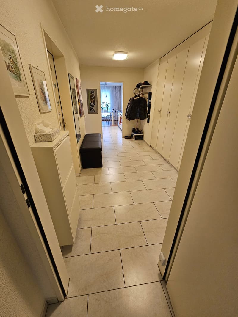 A narrow hallway with tiled flooring, white walls, and several doors. There are framed pictures on the left wall, a small bench, and a coat rack with jackets on the right side. A doorway at the end leads to another room.