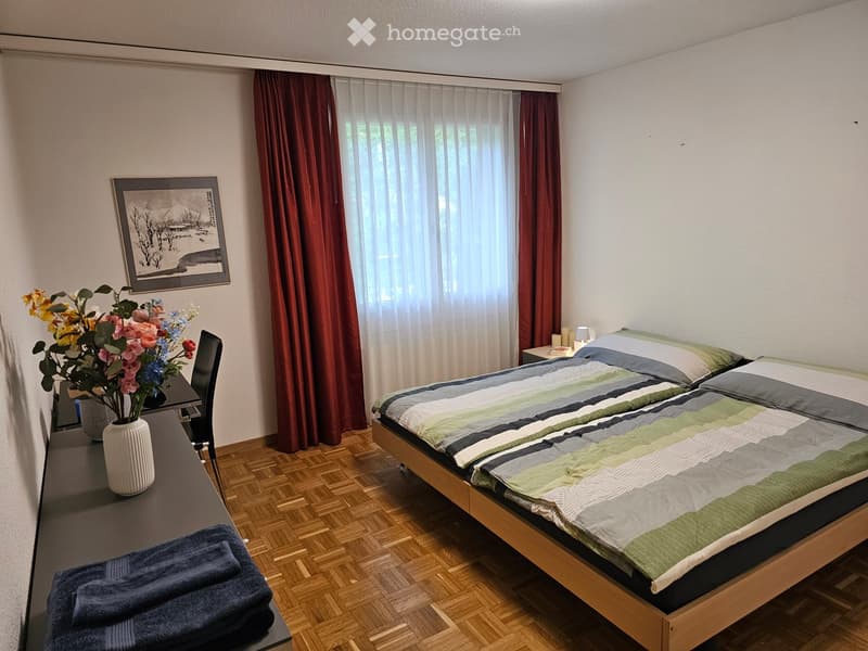 Cozy bedroom with a wooden bed, striped bedding, a small table with a vase of flowers, a chair, and a picture frame on the wall.