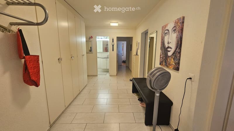 Hallway with tiled floor, white walls, and a fan placed on a small black table. A painting is visible on the right wall, and a red bag hangs on the left side.