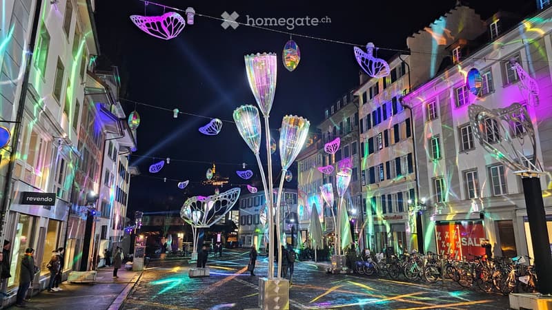 Illuminated street with shops, modern art installations, bicycles parked on the side, pedestrians walking, buildings with modern architectural designs, colorful lighting