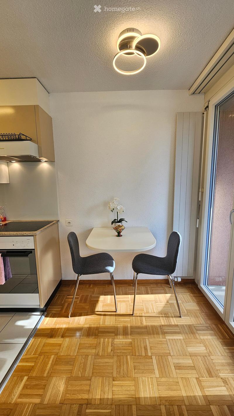 A kitchen-dining area with a modern design and wooden floor. It features a white dining table with two grey chairs, a modern round ceiling light, and a white stove. A kitchen island is situated at the back with a flower vase on it. The kitchen also has a