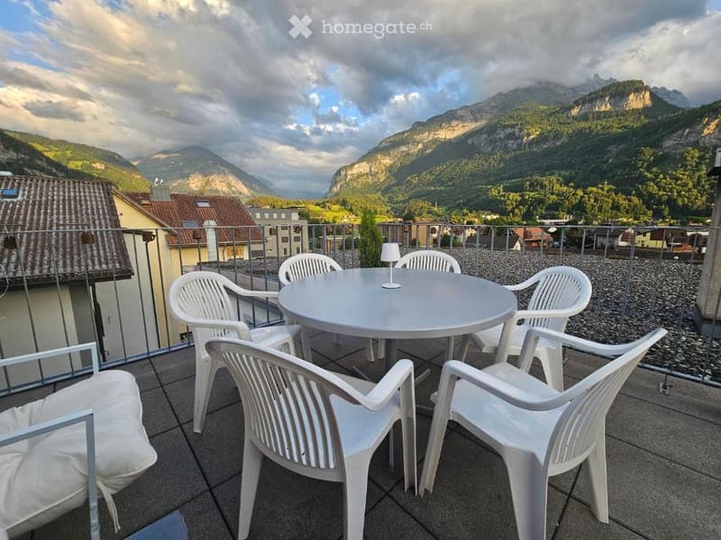 Outdoor terrace with round table, white chairs, metal railings, view of mountains and buildings, white cushion on chair, table lamp.