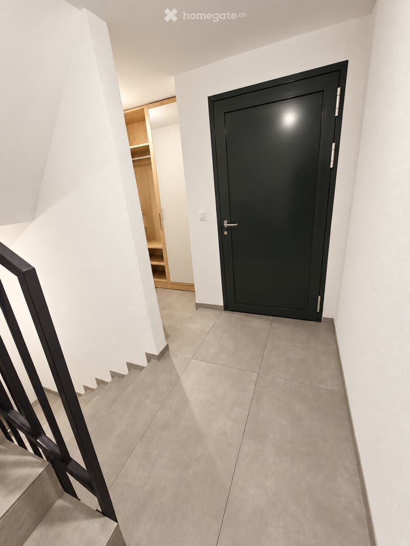 Modern hallway with white walls, tiled floor, black staircase railing, and a closed black door leading to another room.