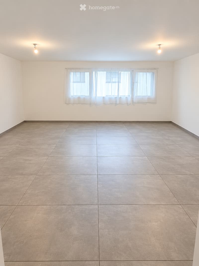 Empty room with tiled floor and white walls, featuring large windows with sheer curtains and two ceiling lights.
