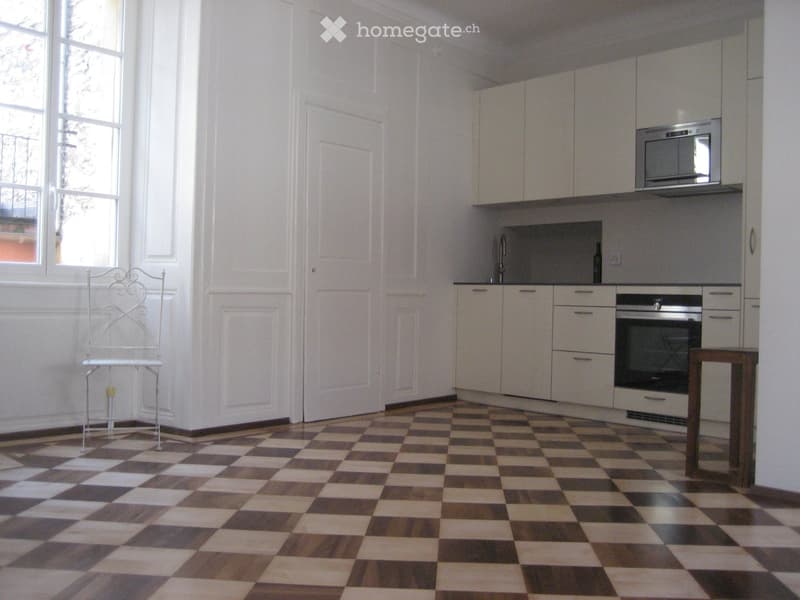 Modern kitchen with white cabinets, stainless steel appliances, and a checkered floor. Features a small wooden table and a white metal chair.