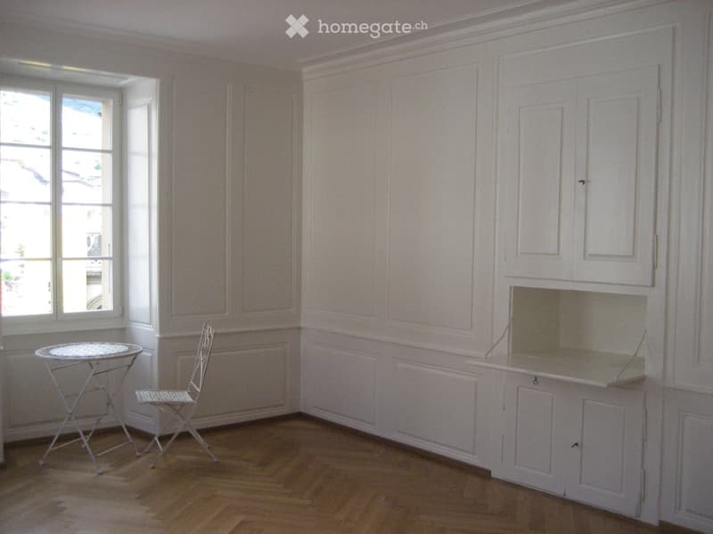 Bright room with wooden flooring, white cabinetry, a small round table, and a matching chair near a large window.
