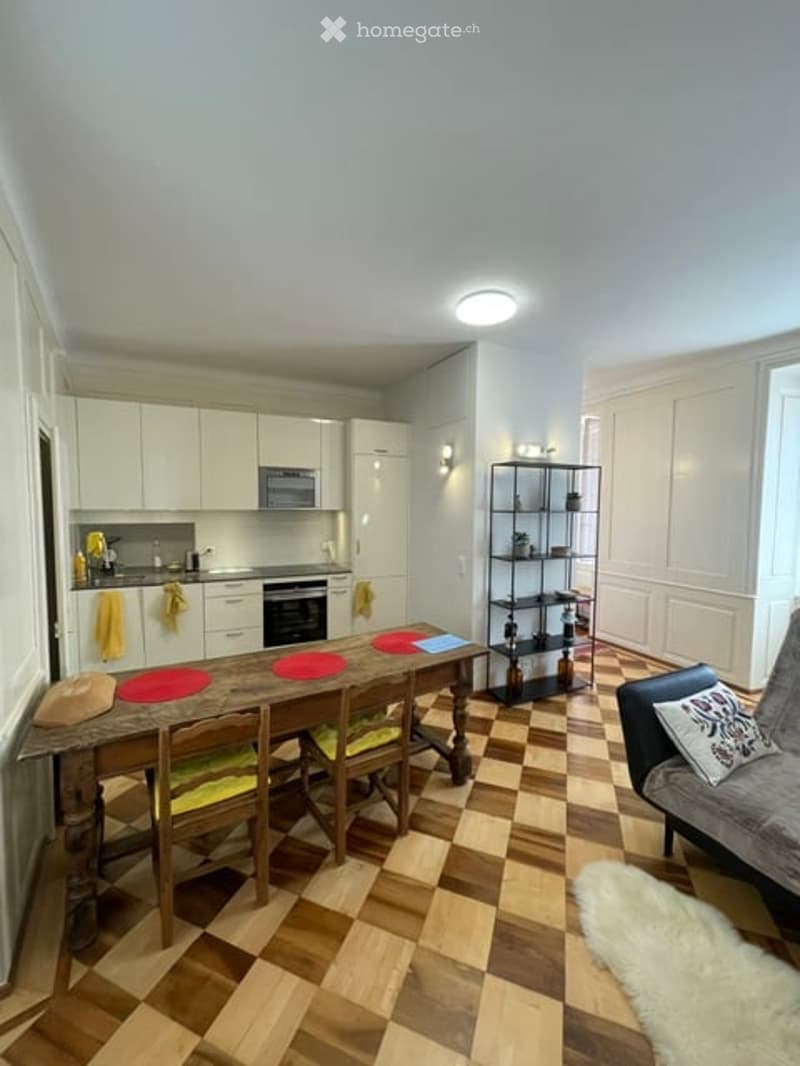 A modern, open-plan kitchen and living area with white cabinets, a wooden dining table, and a comfortable sofa. The space features a checkered floor and a sleek, contemporary design.