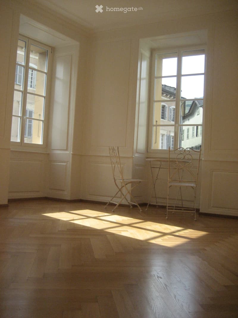 Bright, unfurnished room with wooden flooring, large windows, and two chairs near the windows.