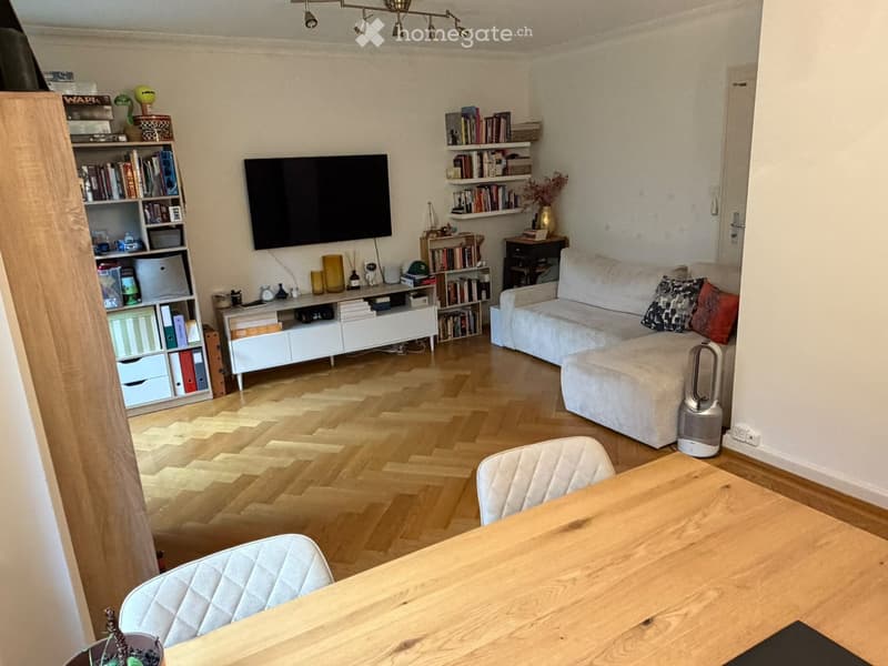 Modern living room with light wooden flooring, a white sofa, a wooden dining table with two chairs, a TV mounted on the wall, and several bookshelves filled with books and decorative items.