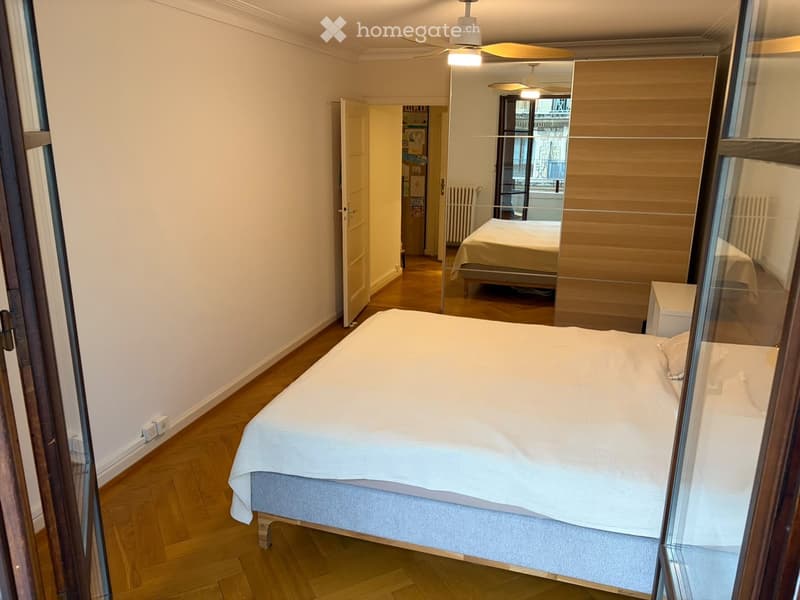 Spacious bedroom with wooden flooring, large bed with white bedding, nightstand, and open door leading to another room.