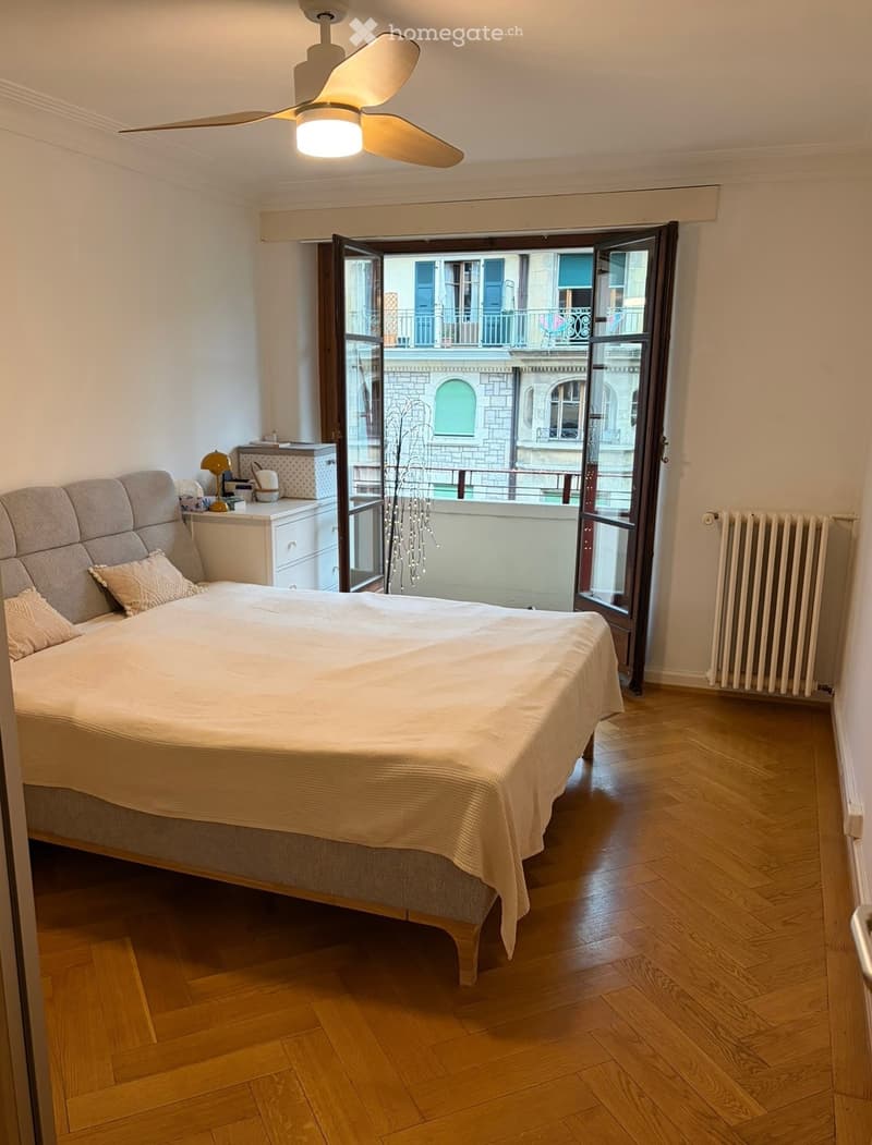 Bedroom with wooden flooring, large bed with pillows and blanket, nightstand with lamp, radiator, and open sliding doors leading to a balcony.