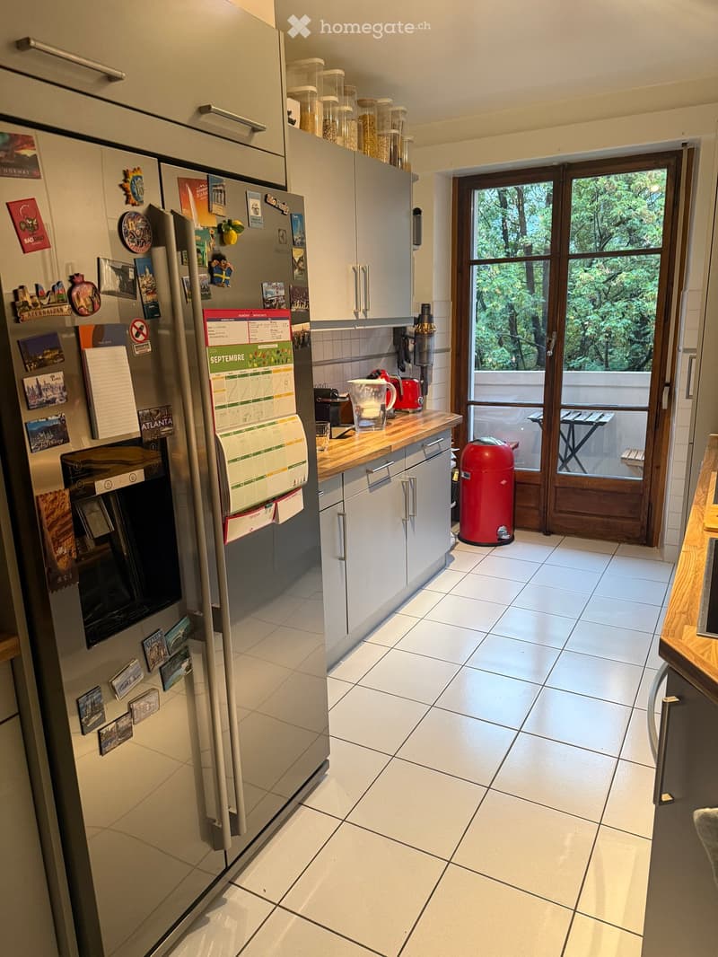 Modern kitchen with white tiled floor, grey cabinets, stainless steel refrigerator adorned with magnets and photos, a wooden countertop with a blender, and a red trash bin near the door leading to a balcony.