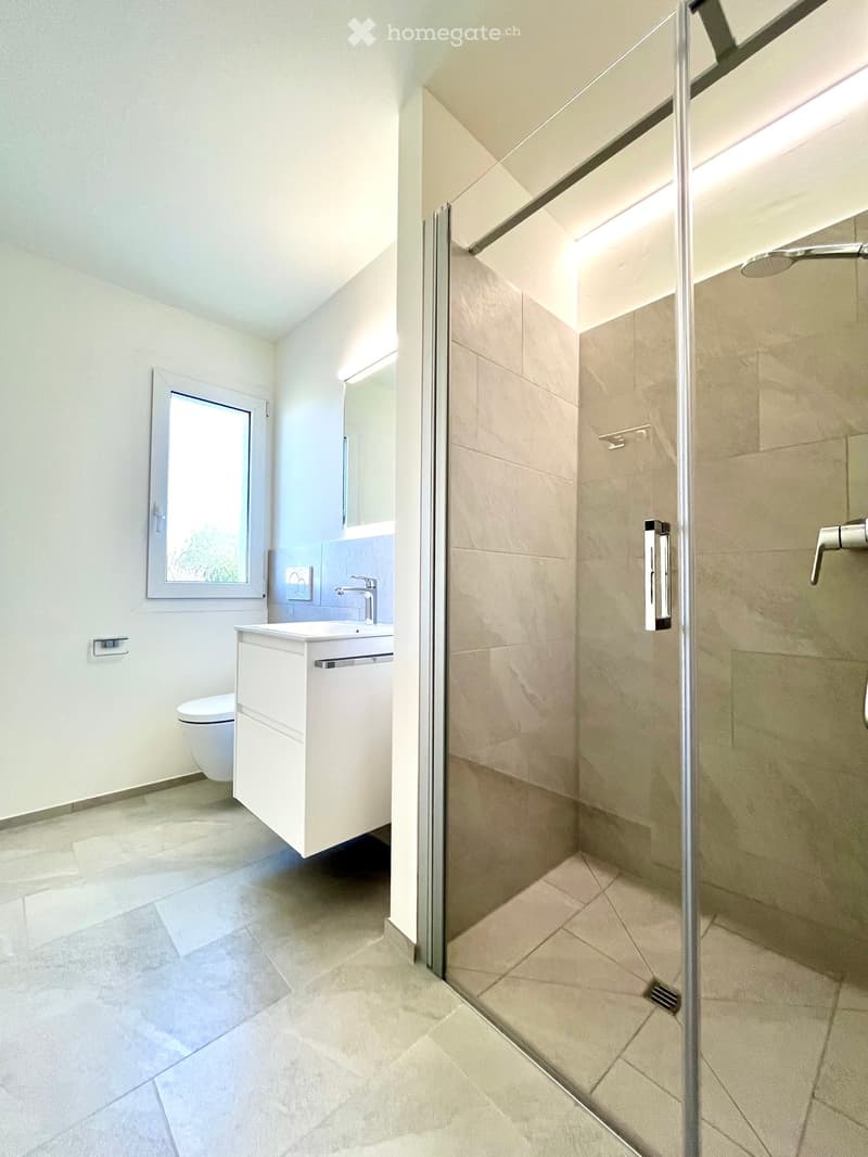 Modern bathroom with white tiled walls, a glass-enclosed shower, a white sink with a chrome faucet, and a toilet. The room features a large window with a view of greenery outside.