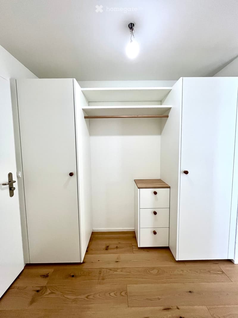Empty room with white walls, wooden floor, built-in closet with shelves, and a small white cabinet with drawers.